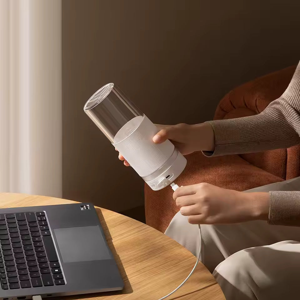 A person plugging a charging cable into the white Alphinea portable blender next to a laptop. Showcasing convenient on-the-go charging for fresh noon break office smoothies.